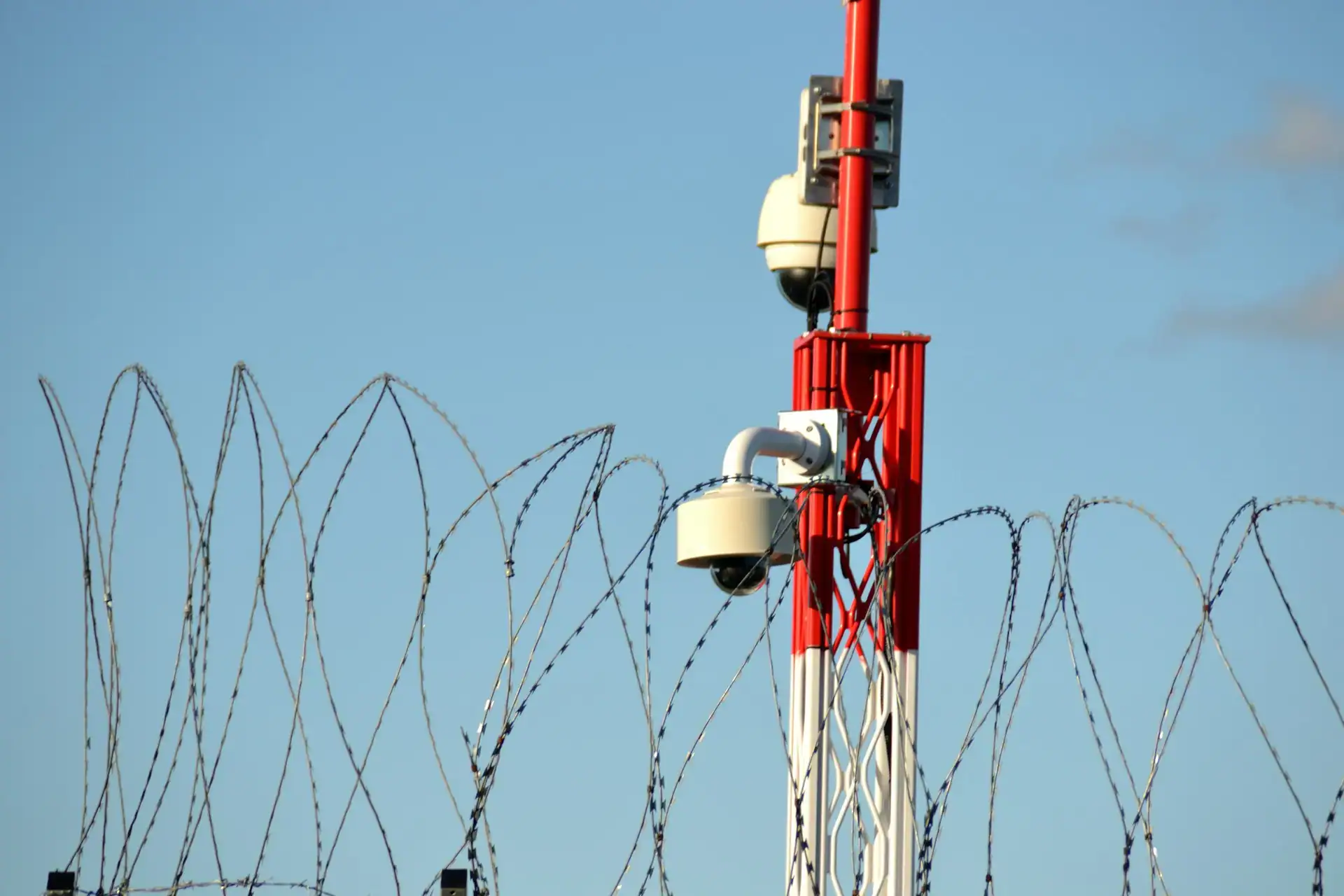 Eine rot-weiß gestreifte Säule mit zwei weißen Dome-Sicherheitskameras hinter einer Barriere aus spiralförmigem S-Draht (Natodraht) vor blauem Himmel